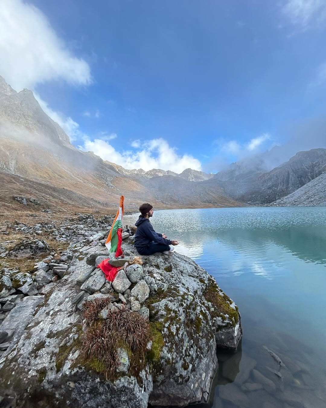 Nirvaan Birla meditating by a mountain lake with a saffron flag beside him, under a bright blue sky.