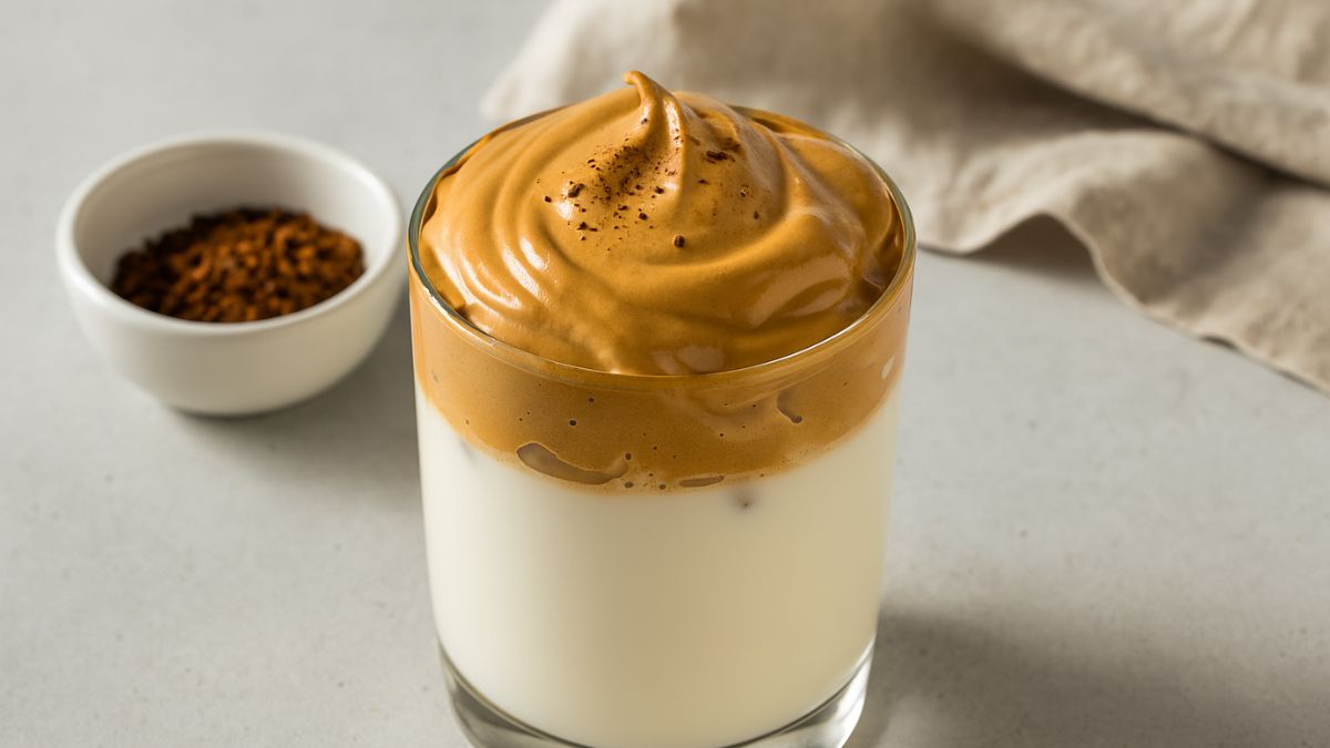 A high-resolution digital photograph showcases a glass of dalgona coffee with whipped coffee foam atop iced milk, next to a small bowl of instant coffee and a soft beige napkin.
