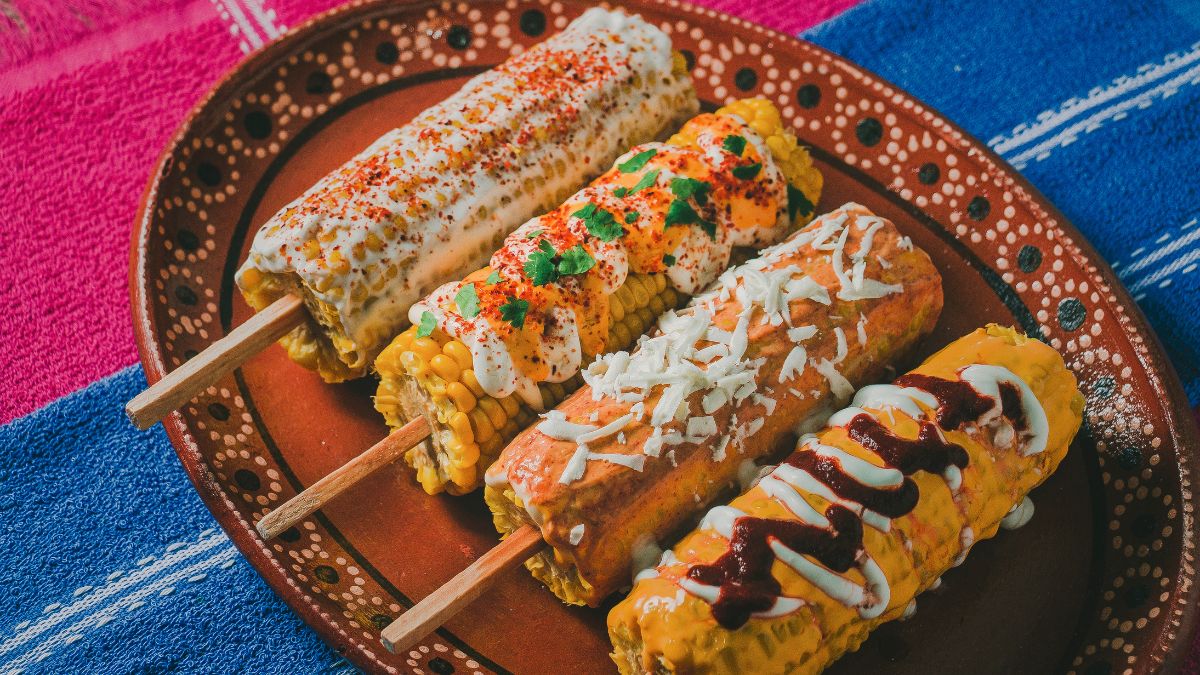 Four Mexican-style corn on the cob sticks on a rustic plate with colorful toppings