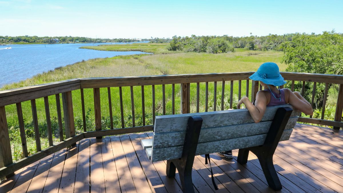 Woman in a blue hat sitting on a bench overlooking a river and wetlands in Florida