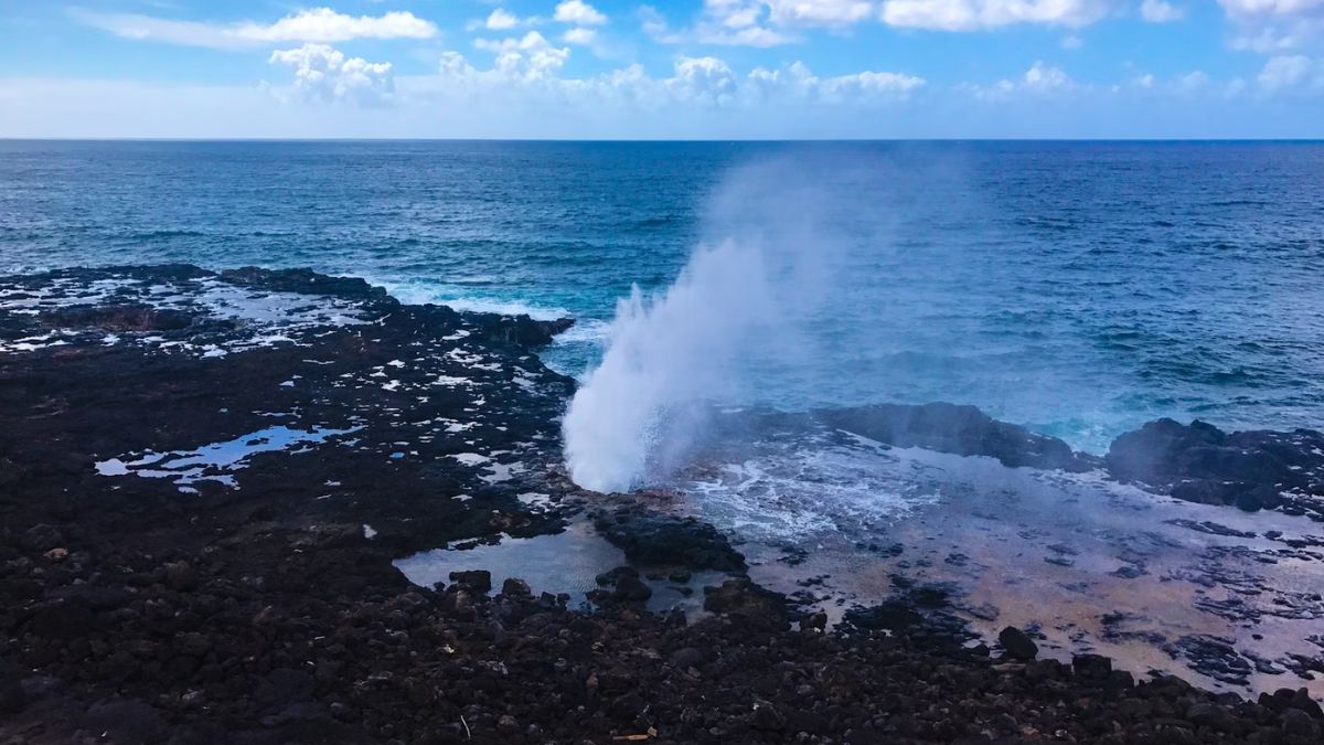 Ocean spray shoots through volcanic rocks at a Hawaiian coastline under a blue sky