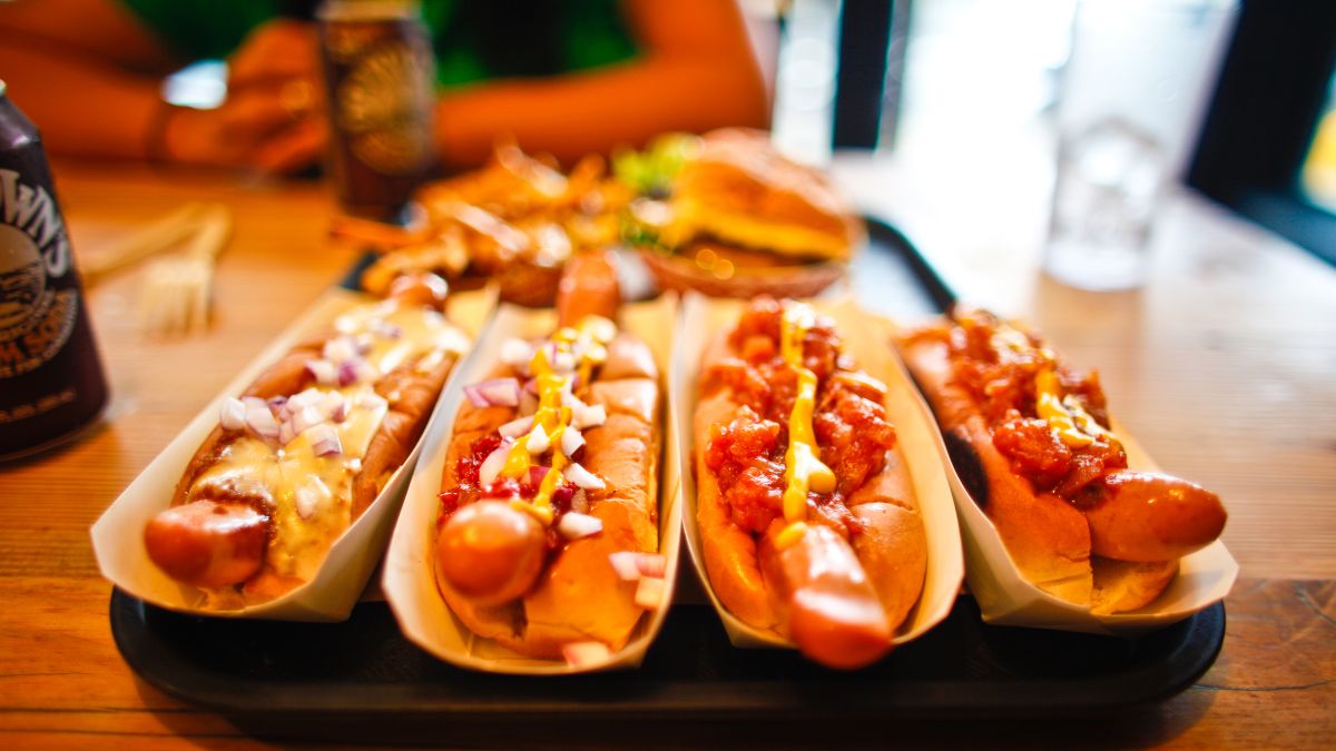 A tray of five loaded hot dogs with various toppings including cheese, onions, and mustard, placed on a wooden table at a restaurant