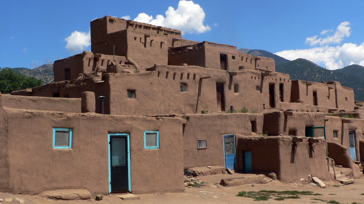 Adobe structures of Taos Pueblo under a bright sky, with turquoise-painted doors and mountains in the background