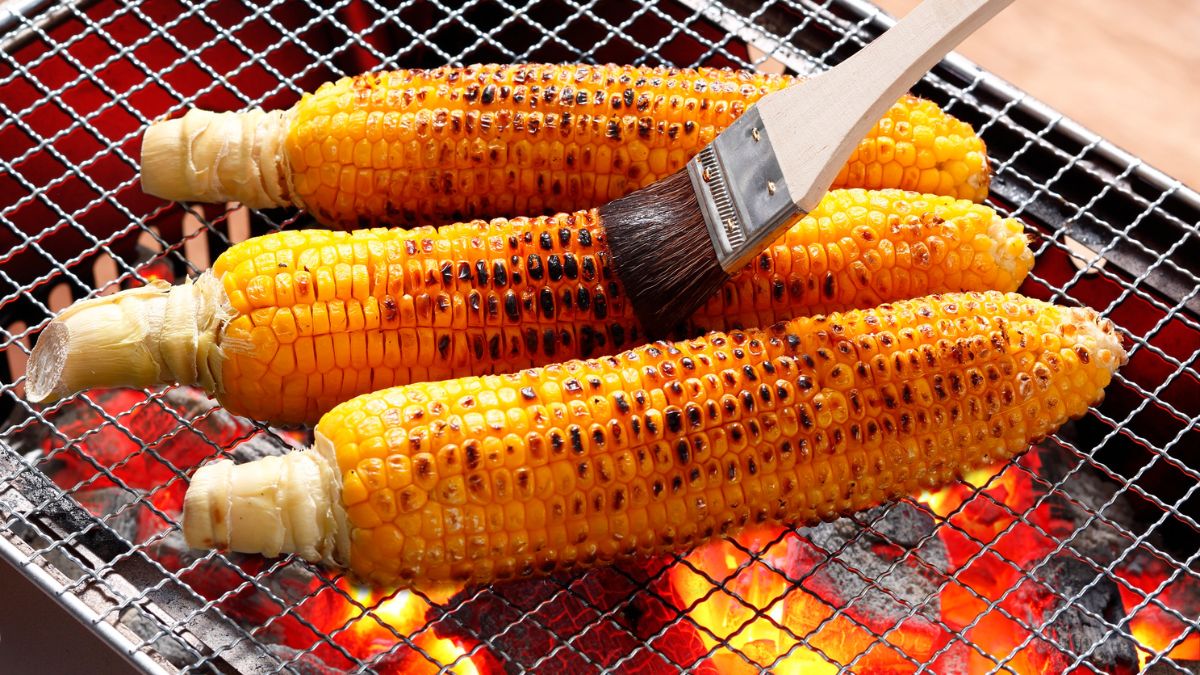 Grilled corn on the cob being brushed with butter over hot charcoal